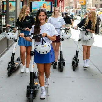 Four riders walking Ride by iPakket e-scooters along a Manhattan sidewalk